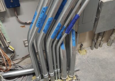 Metal electrical conduits organized and labeled with blue tape. They are mounted on a wall and secured at the bottom with yellow insulation material. Electrical panel and wires are visible in the background.