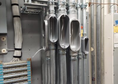 A series of metal conduits bending upward with open junction boxes attached, revealing exposed wiring inside. Nearby are cable bundles and a mounted panel with fuses.