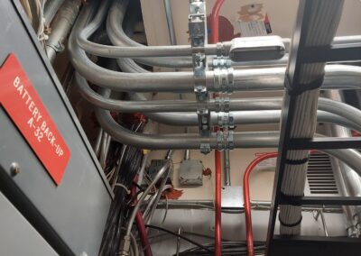 View of an electrical system with multiple conduits, wiring, and metal fasteners. A sign labeled "Battery Back-Up A52" is visible on the left.