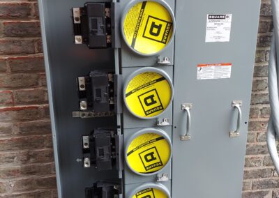 An open electrical panel with four circuit breaker switches exposed, mounted on a brick wall. The panel is labeled 'Square D' and features yellow safety covers on the breakers.