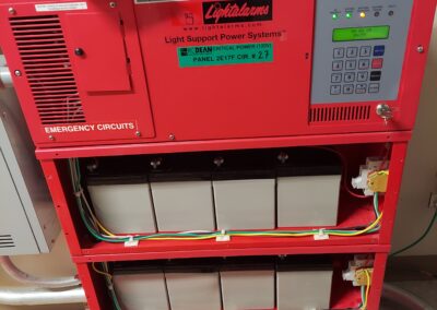 Red electrical panel labeled "Light Support Power Systems" with emergency circuits. Below are three rows of batteries connected with cables. Keypad and display screen are on the right side of the panel.