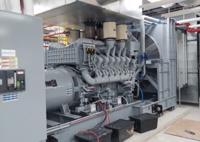 A large industrial generator with multiple connected components is installed in a control room, surrounded by ventilation ducts, electrical panels, and several buckets on the floor.