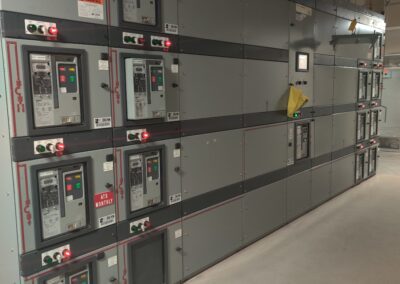 A row of industrial electrical control panels with various buttons, switches, and meters in a utility room. Warning labels and indicators are visible on the panels.