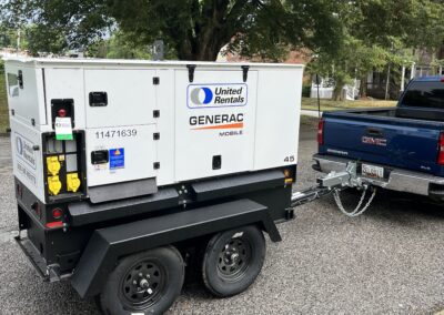 A Generac mobile generator from United Rentals is attached to the back of a dark-colored pickup truck, parked on a paved road with trees in the background.