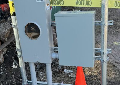 Electrical boxes mounted on poles at a construction site with a caution tape and an orange traffic cone in front. Graffiti is visible on a wall in the background, and people are walking on a path.