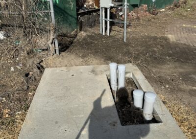 Concrete slab with piping and utility connections, located near a fence covered in graffiti. Shadow of person visible in foreground.