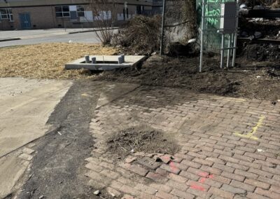 A brick pathway transitions to a paved surface, with construction equipment and barriers nearby. In the background, there's a single-story building, leafless trees, and scattered patches of grass.