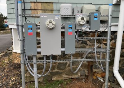 Three electrical meter boxes attached to the exterior of a building with light green shingles. Conduits and cables connect the meters to the house, with a stone foundation visible.