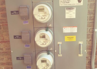 Electrical panel with multiple meters and switches. Label warns of hazardous voltage. Panel doors are secured with padlocks. Brick wall in the background.