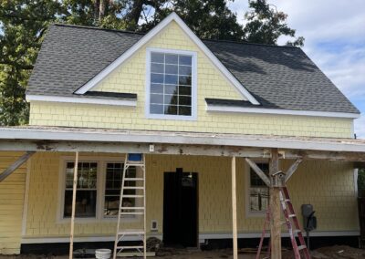 A yellow house with a gray roof is under construction with ladders and building materials in front. There is scaffolding set up on the porch area and a large tree in the background.