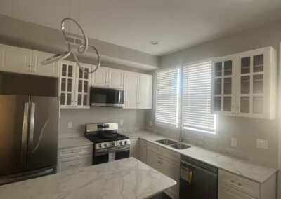 A modern kitchen with white cabinetry, marble countertops, stainless steel appliances, and a center island under a unique pendant light fixture. There's a large window near the sink.