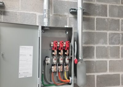 An open electrical panel with various colored wires and exposed connections mounted on a cinder block wall. Conduit pipes are connected to the panel from above and beside.